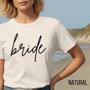 Puede incluir: Una camiseta blanca con la palabra "bride" escrita en cursiva negra. La camiseta la lleva puesta una persona de pie en una playa.