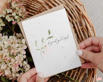 May include: A white seed paper greeting card with a textured surface featuring a green botanical illustration of a leafy stem with small pink buds and the black cursive text beyond grateful, paired with a brown envelope.