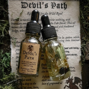 May include: Two glass dropper bottles with black caps, labelled "Devil's Path," rest on a printed parchment with text and a dragon illustration. The bottles are surrounded by green moss and dried plants. One bottle has a brown label.