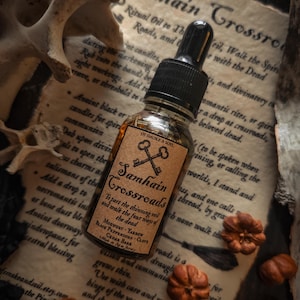 May include: A bottle of Samhain Crossroads ritual oil with a black dropper, on vintage paper with text. The label has keys and text. Dried gourds and a bone are in the background.