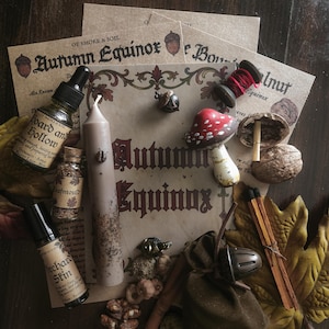 Peut inclure: Une collection d'objets comprenant une bougie effilée beige, des flacons en verre avec des étiquettes Orchard Skin et Leafmould, une figurine de champignon rouge et blanc, une pochette en tissu marron, des noix, une bobine de fil rouge et des cartes Autumn Equinox.