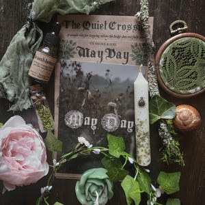 May include: An assortment of mystical items arranged on a dark wooden surface. Items include a candle, a small bottle, a book with the text "May Day", a pink peony, and a green rose-shaped candle. Decorative greenery and a snail shell are also present.
