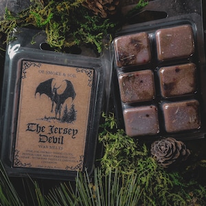 May include: A six-pack of wax melts in a clear plastic package. The wax melts are a dark brown color and have a speckled appearance. The package has a label that reads "The Jersey Devil Wax Melts" and lists the scents: pine, fir, smoked wood, clove, peppercorn, burnt marshmallow, cedar, pine needles, oakmoss, and cinnamon.