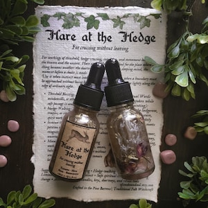 May include: Two small glass dropper bottles with black caps resting on a piece of textured paper. The left bottle features a label with a hare illustration and the text "Hare at the Hedge". The right bottle contains visible dried botanicals.