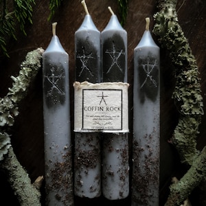 May include: Four grey taper candles featuring a central paper label with the text COFFIN ROCK and For old stones, old stories, and the dead that remember OF SMOKE & SOIL. Each candle has a dark grey painted symbol and a textured, soil-like base.