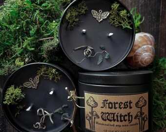 Könnte beinhalten: Schwarze Sojawachskerzen in Dosen, verziert mit Moos, Metallanhängern und getrockneten Pflanzen. Eine Dose trägt die Aufschrift "Forest Witch" und "Handcrafted soy wax candle". Die Kerzen sind von Moos und Schneckenhäusern umgeben.