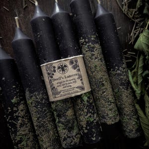 May include: A collection of black candles with a textured, herbal coating. A paper label wrapped around the center of the candles reads "Devil's Lantern" with additional text and a graphic. The candles are arranged on a dark wooden surface.