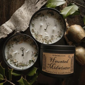 Peut inclure: Trois bougies en boîte noire avec de la cire blanche, des herbes décoratives et un charme de clé. Une bougie porte une étiquette indiquant "Haunted Midwinter". Les bougies sont sur une surface en bois sombre avec de la verdure.