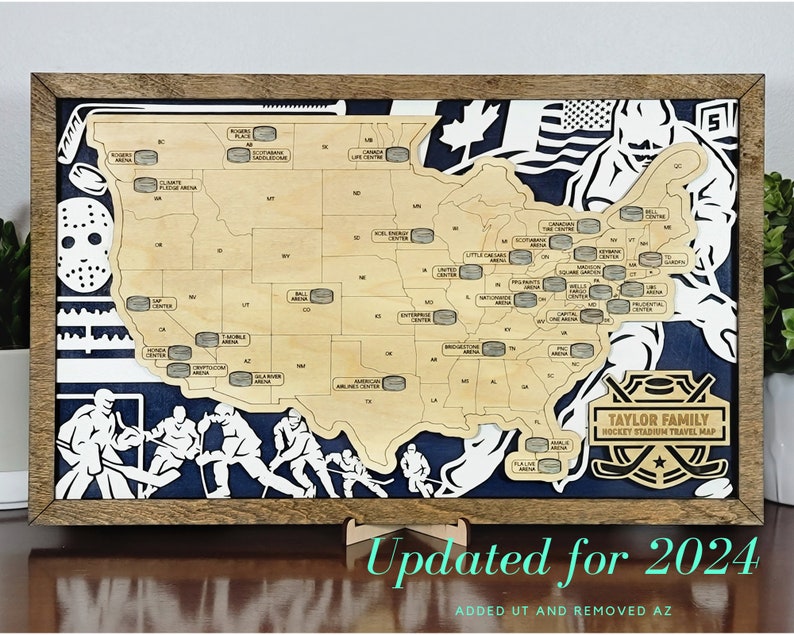 May include: A wooden map of the United States with hockey arenas marked on it. The map is framed in a brown wood frame and has a blue background. The text "TAYLOR FAMILY HOCKEY STADIUM TRAVEL MAP" is printed on the map. The text "Updated for 2024 ADDED UT AND REMOVED AZ" is printed below the map.