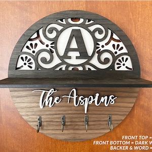 May include: A wooden key holder with a shelf and five hooks. The top of the key holder is a half-circle with a decorative design and the letter "A" in the center. The bottom of the key holder is a rectangular shelf with the text "The Aspins" in white lettering. The key holder is made of ebony, dark walnut, and white wood.