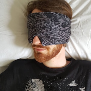 May include: A person wearing a black t-shirt with a Star Wars graphic print, lying in bed with a gray and black patterned sleep mask over their eyes.