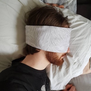 May include: A person sleeping in bed with a white and gray patterned sleep mask over their eyes.