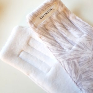 May include: A pair of white fleece gloves with a brown and white patterned design on one glove. The patterned glove has a white label with the text "ILKE USLUGA" on it.