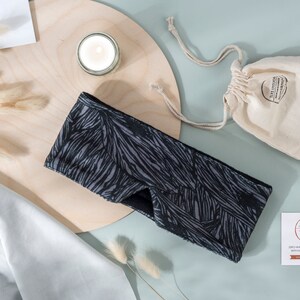 May include: Black and gray sleep mask with a geometric pattern. The mask is on a wooden surface with a candle and a small white bag.