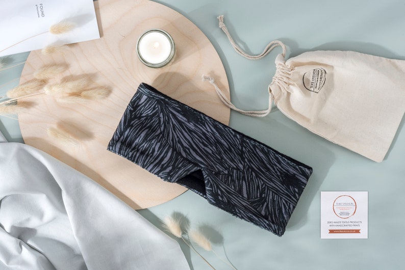 May include: Black and gray sleep mask with a geometric pattern. The mask is on a wooden surface with a candle and a small white bag.