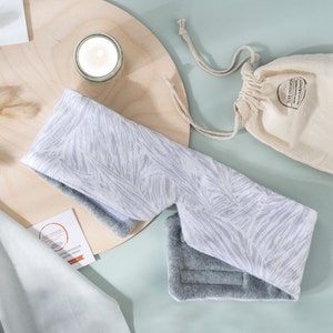 May include: A white and gray eye mask with a zebra print design. The eye mask is made of soft fabric and has a gray fleece lining. The eye mask is on a wooden surface with a small white candle and a white drawstring bag.