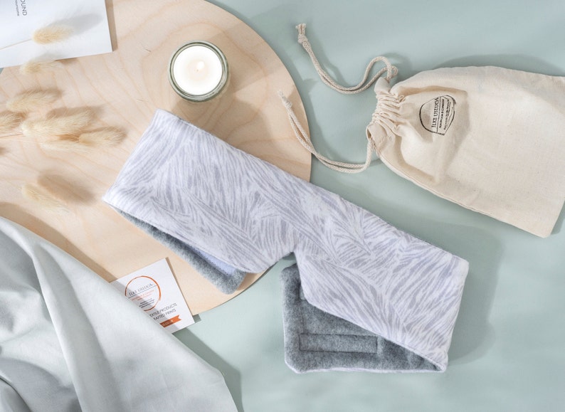 May include: A white and gray eye mask with a zebra print design. The eye mask is made of soft fabric and has a gray fleece lining. The eye mask is on a wooden surface with a small white candle and a white drawstring bag.