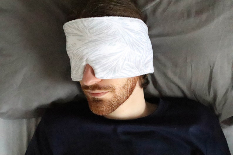 May include: A person wearing a white sleep mask with a leaf pattern on a gray pillow.