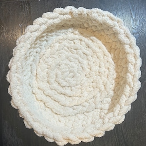 May include: A round, off-white basket crafted from thick, chunky yarn. The basket features a circular base and raised, textured walls created by a continuous, spiraling crochet stitch pattern that gives the object a soft, plush appearance.