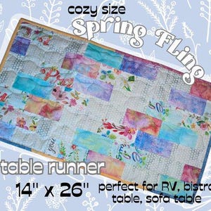 May include: A pastel-colored quilted table runner with a patchwork design. The runner is 14 inches by 26 inches and features the text "Spring Fling" and "table runner".