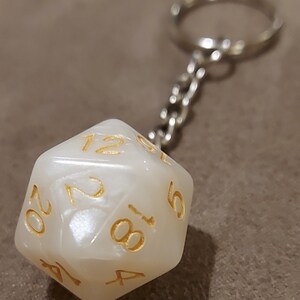 May include: A white, iridescent, 20-sided dice keychain with gold numbers. The keychain is attached to a silver metal chain.