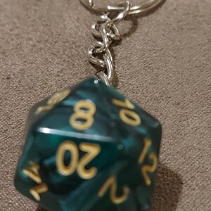May include: A teal green 20-sided die keychain with gold numbers. The die is attached to a silver keychain.