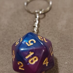 May include: A purple and blue iridescent d20 dice keychain with gold numbers. The keychain is attached to a silver metal ring.