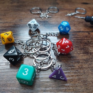 Puede incluir: Un juego de seis llaveros de dados coloridos, cada uno con un número diferente, unidos a una cadena plateada con múltiples llaveros. Los dados son rojo, azul, amarillo, negro, verde y morado.