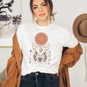 May include: White t-shirt with a graphic design of a bull skull, sun, and moon with the words "WILD SOUL" on either side of the skull.