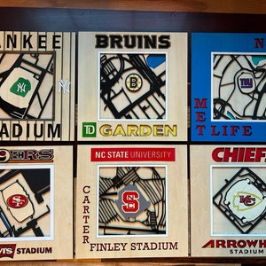 May include: Six wooden plaques with team logos and stadium names. The plaques are arranged in two rows of three. The top row features the New York Yankees, Boston Bruins, and New York Giants. The bottom row features the San Francisco 49ers, NC State University, and the Kansas City Chiefs.