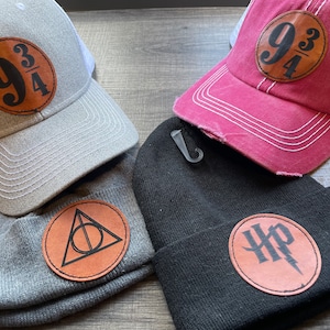 May include: Four hats and beanies with leather patches. Two baseball caps have the "9 3/4" logo, one in gray and one in maroon. A gray beanie has a Deathly Hallows symbol, and a black beanie has the "HP" lightning bolt logo.