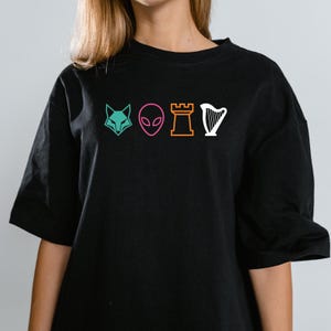 Peut inclure: T-shirt noir avec une tête de renard turquoise, une tête d'extraterrestre rose, une pièce d'échecs de tour orange et une harpe blanche. Le graphisme est de style linéaire. Le t-shirt a des manches courtes et un col rond.