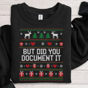 Peut inclure: Sweat-shirt noir avec un motif pixelisé de Noël. Le motif comprend des rennes, des sapins, des cœurs, des flocons de neige et des boîtes cadeaux rouges, vertes et blanches. Le texte "BUT DID YOU DOCUMENT IT" est en blanc.