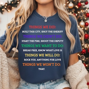 K&ouml;nnte beinhalten: Marineblaues T-Shirt mit mehrfarbigem Text. Der Text listet Aussagen wie "BAUT DIESE STADT", "ERSCHOSS DEN SHERIFF" und "ROCK YOU" auf. Ein auff&auml;lliges Kleidungsst&uuml;ck f&uuml;r den Alltag.