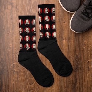Puede incluir: Calcetines negros con retratos de figuras históricas en círculos rojos. Los calcetines están sobre una superficie de madera, con un par de zapatillas grises en el fondo. Accesorio único con varias personas.