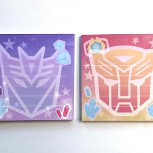 Può includere: Due blocchi per appunti quadrati con simboli dei Transformers. Uno è viola con un simbolo bianco, stelle e forme cristalline. L'altro è rosa e giallo con un simbolo dorato, stelle e forme cristalline. Entrambi hanno aree a righe.