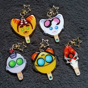 May include: Five colorful character keychains shaped like popsicles. Each keychain features a different character with unique facial features and colors. The keychains are made of clear acrylic and have a gold star charm attached to the top.