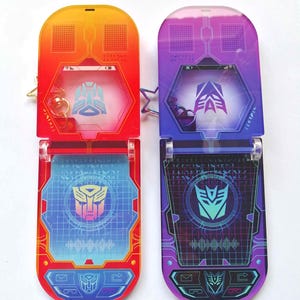 Puede incluir: Dos colgantes de teléfono de acrílico con degradados rojo-naranja y morado-azul, cada uno con un logotipo de Transformers y varios diseños de pantalla. El texto "Flipped open" y "different screen shapes" son visibles.
