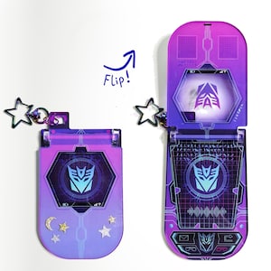 Puede incluir: Un teléfono plegable morado y rosa con un logotipo de Transformers. El teléfono está abierto, revelando una pantalla con el mismo logotipo. Un dije en forma de estrella cuelga del teléfono. La palabra "Flip!" está escrita encima.