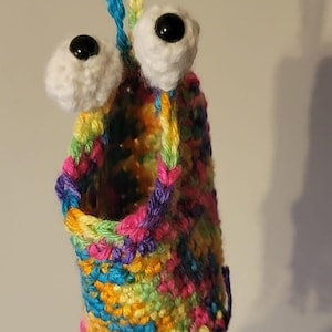 May include: A colorful crocheted pouch with a rainbow pattern and two white googly eyes with black pupils. The pouch has a long, colorful crocheted cord for hanging.