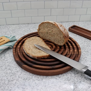 May include: A round, dark wood bread board with concentric grooves holds a sliced loaf of bread and a stainless steel knife. A second loaf of bread sits next to the slice. A wooden serving tray and a striped towel are also visible.