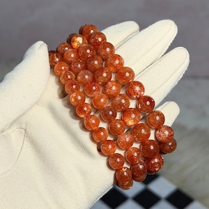 Può includere: Un bracciale a più fili composto da perline rotonde e levigate in pietra del sole. Le perline presentano una calda tonalità arancione-marrone traslucida con inclusioni scintillanti interne che catturano la luce, creando un effetto riflettente e brillante.