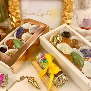 May include: Two boxes of assorted crystals, one gold and one white, with a clear window showing the crystals inside. The boxes are surrounded by other crystals, a smudge stick, and a pendulum.