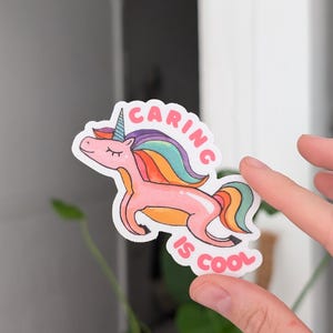 May include: A sticker featuring a pink unicorn with a rainbow mane and horn. The text "Caring is Cool" is written above and below the unicorn in a playful font. The sticker is held by a hand.