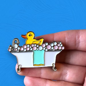 May include: A yellow rubber ducky in a white bathtub with pink and white bubbles. The bathtub has a blue towel and gold accents.