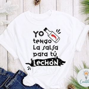 May include: White t-shirt with black text that reads "Yo tengo la salsa para t&uacute; Lech&oacute;n" with a black and white illustration of a pig and a red chili pepper in a bottle.
