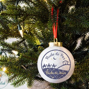 May include: A white Christmas ornament with a blue and gold design. The ornament features the text "Noche de Paz" and "Noche de Amor" with a silhouette of the three wise men riding camels and a star.
