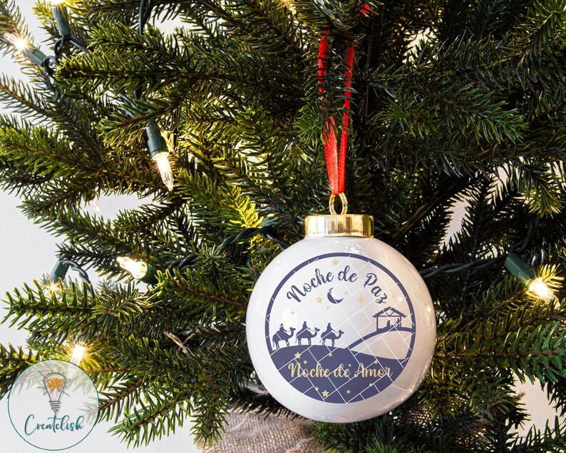 May include: A white Christmas ornament with a blue and gold design. The ornament features the text "Noche de Paz" and "Noche de Amor" with a silhouette of the three wise men riding camels and a star.