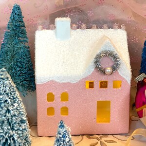 K&ouml;nnte beinhalten: Ein rosa, glitzerbedecktes Keramikhaus mit schneewei&szlig;em Dach und Schornstein, verziert mit einem silbernen Kranz und einer Perle. Miniatur blaue Flaschenb&uuml;rstenb&auml;ume und Weihnachtsschmuck umgeben das Haus und schaffen eine festliche Urlaubsszene.