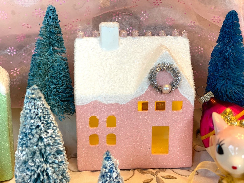 K&ouml;nnte beinhalten: Ein rosa, glitzerbedecktes Keramikhaus mit schneewei&szlig;em Dach und Schornstein, verziert mit einem silbernen Kranz und einer Perle. Miniatur blaue Flaschenb&uuml;rstenb&auml;ume und Weihnachtsschmuck umgeben das Haus und schaffen eine festliche Urlaubsszene.